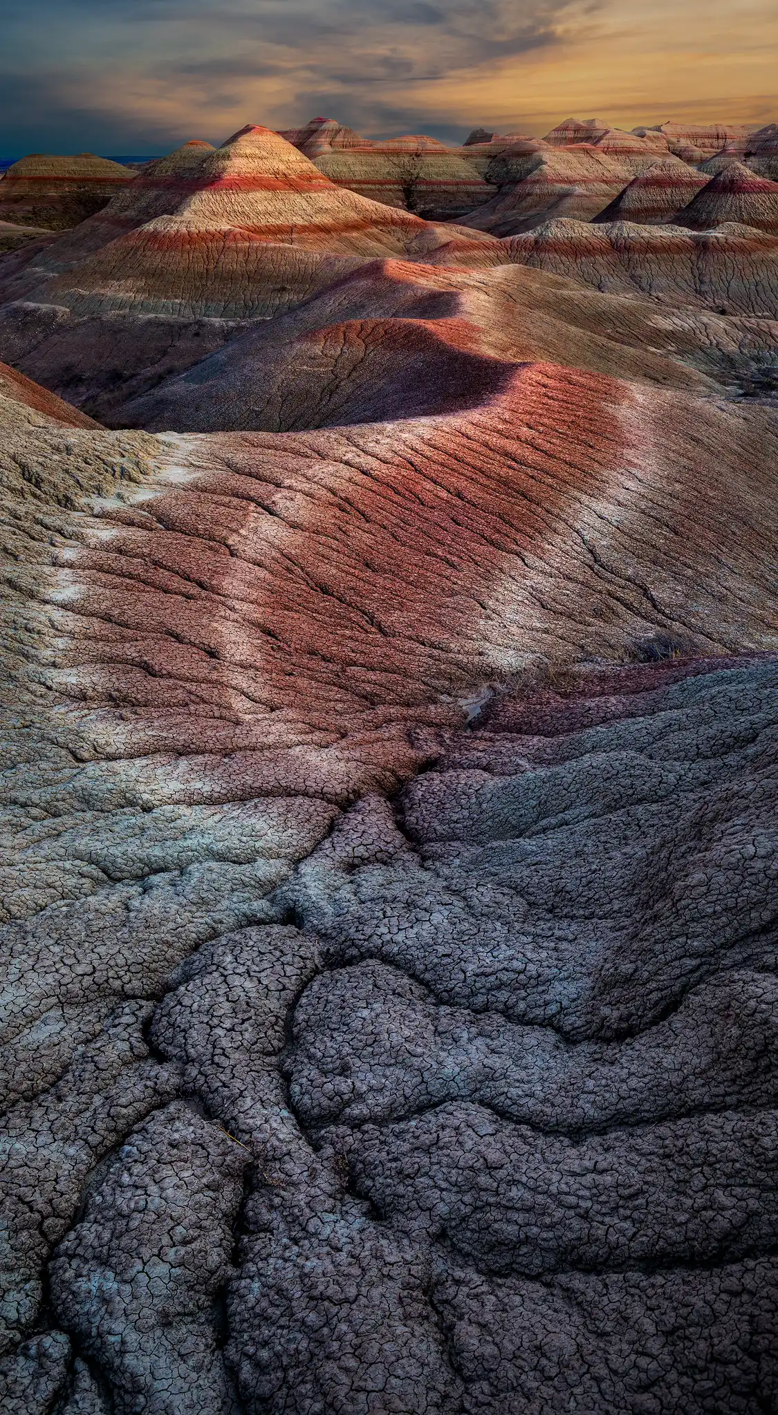 Cracked clay patterns leading toward colorful striped buttes in Badlands National Park during warm evening light.