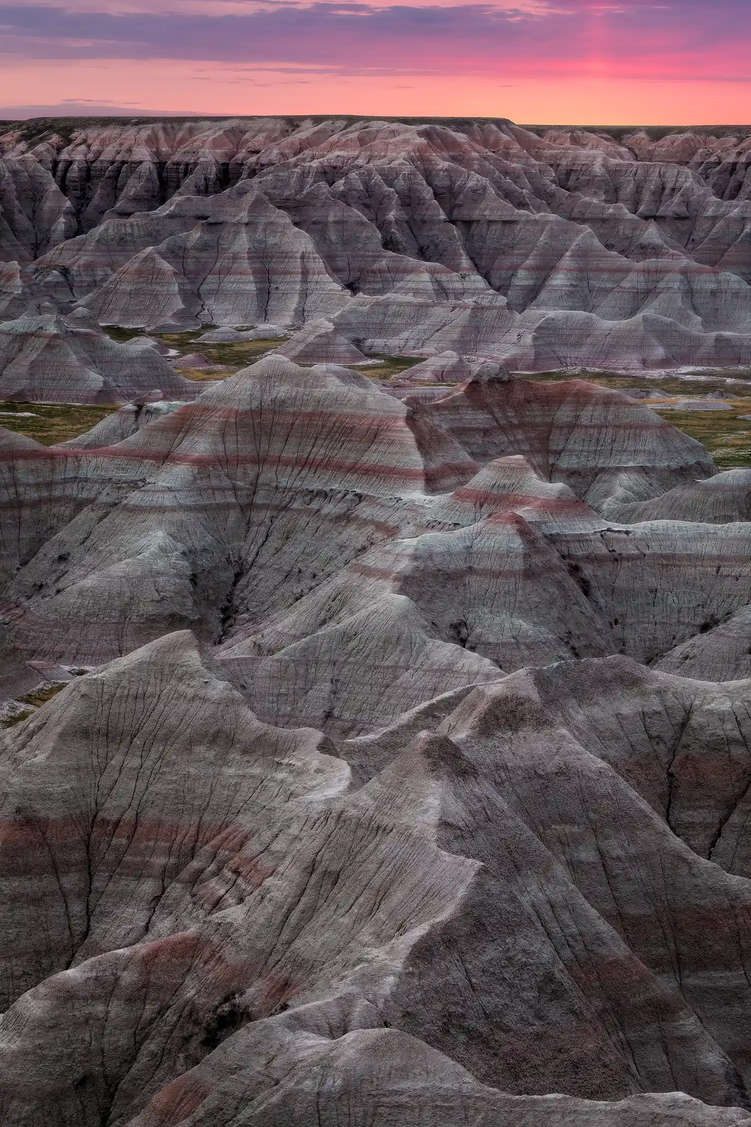 Pink dawn sky over eroded canyons with red and gray rock layers in Badlands National Park.