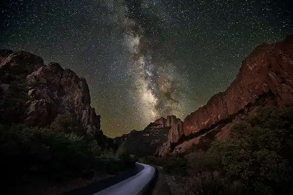 One of our popular workshop locations; Night photo of Chisos Basin Road curving between dramatic cliffs and leading the eye to a distant mountain. The galactic core of the Milky Way sits just above the mountain and rises into the dark sky above Big Bend National Park, Texas.
