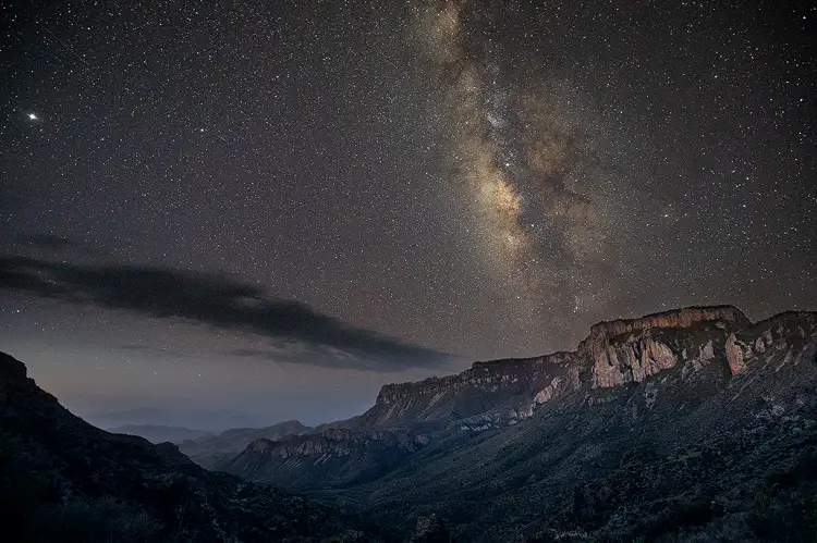 Workshop location; Two thirds of this image is the night sky with the milky way cutting through at a 340 degree angle. The lower portion shows a view across a valley to a rocky ridge that slopes down to the left out of the Chisos Mountains to the hazy plains of Big Bend National Park.