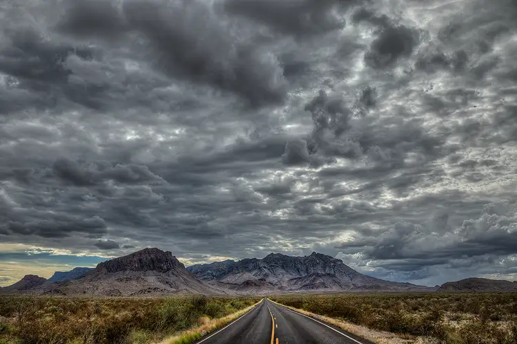 Workshop location; Dark, foreboding clouds fill the top three quarters of this image. The bottom shows a road entering at the center of the frame, the yellow striped, black pavement cutting through a bushy desert. The road leads straight back, a long way, narrowing to a point at the steeply rising Chisos Mountains in Big Bend National Park.