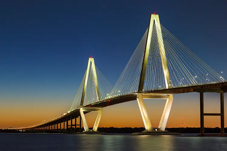 Photography location, Charleston SC; Twilight photo of the Arthur Ravenel Jr. bridge. The sky above is clear and blue, transitioning to an orange glow on the horizon. However it's dark enough that all of the bridge's lights are on, illuminating it's piers and supports making it stand out dramatically against the fading sky.