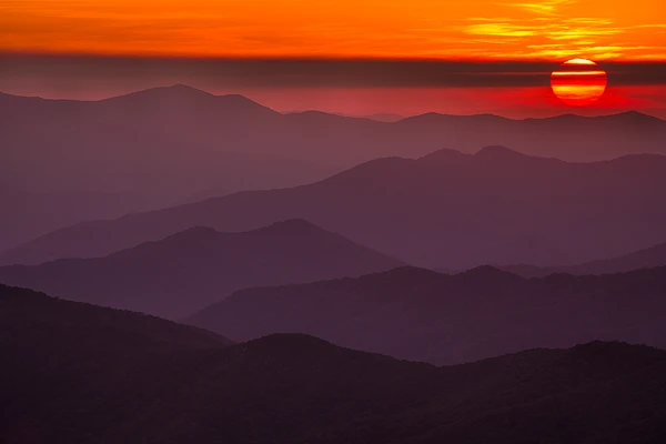Telephoto image of a red-orange sunset partially obscured by striated clouds. Below, in the majority of the image, are the famous, hazy, layered mountains. Great Smoky Mountains National Park continues to be one of the best places to attend a photography workshop.