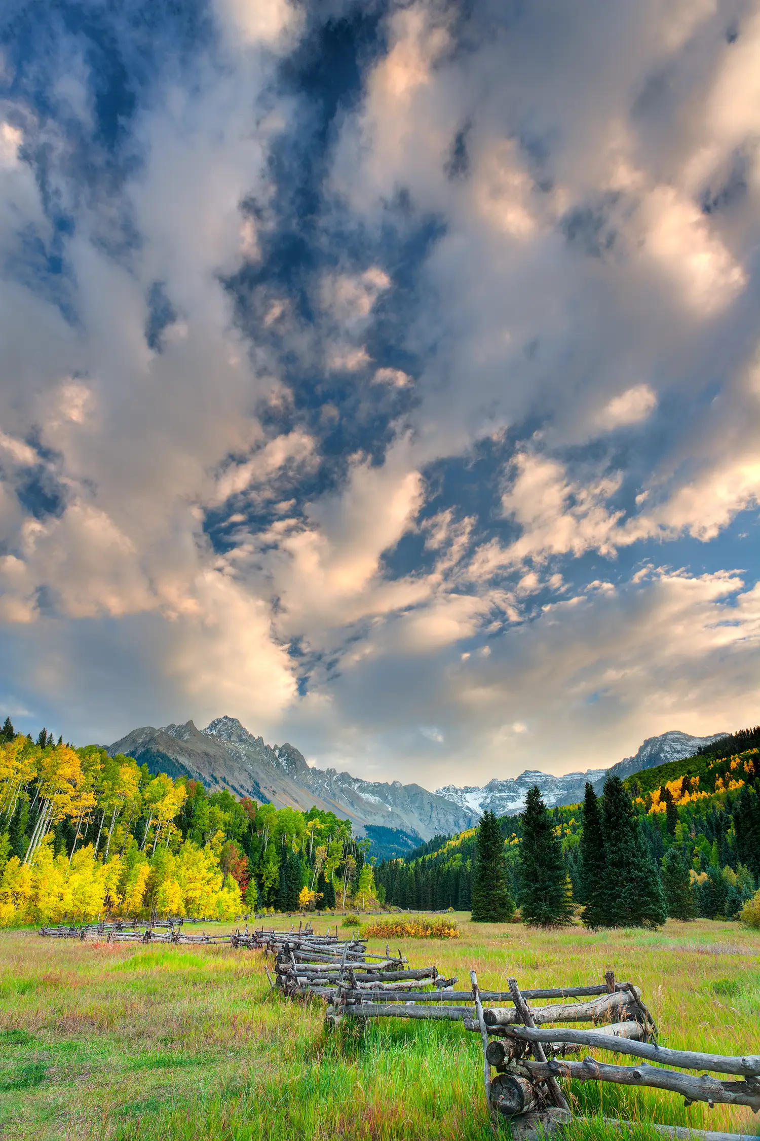 A weathered zigzag fence leading towards a vibrant aspen grove and the majestic Mount Sneffels under a dramatic, cloud-filled autumn sky in Colorado.