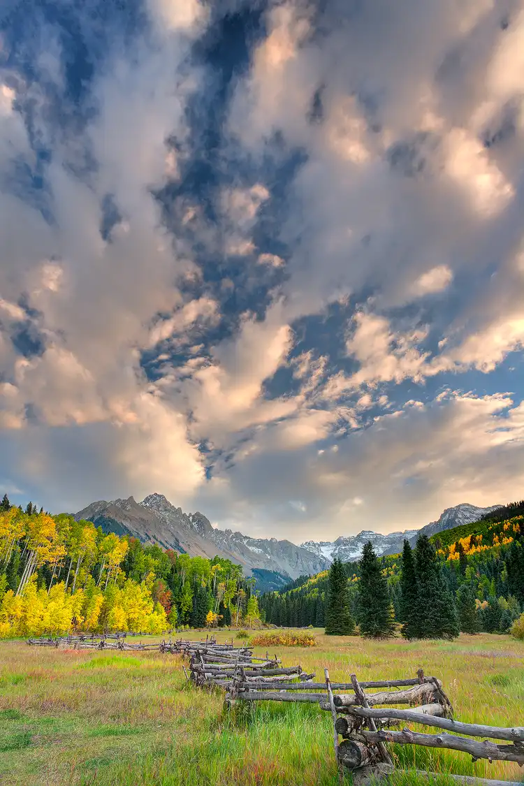 Photography location; The bottom third of this vertical photograph is an old, zig zag fence leading through an autumn field to a colorful stand of aspen trees with Mt. Sneffels above. The top two thirds of the image shows a dramatic arrangement of clouds that are side lit by the evening sun, and due to their long shape, direct attention to the summit of Mount Sneffels.