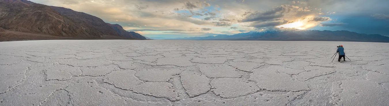 Workshop location; In this panorama, on the right side, a lone photographer in a blue jacket kneels at his tripod on the expansive, white salt flat in Badwater Basin. In the background are mountains and a beautifully clouded sky beginning to glow with sunset.