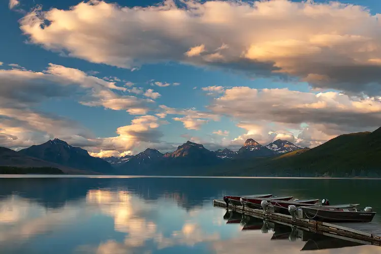 Workshop location; A boat dock, criss crossed with small row boats, juts into the calm lake from the bottom right corner of the frame. Above, warm sunset colors dance on a patchwork of clouds, while in the middle of the frame, looming back in the distance are the mountains of Glacier National Park.
