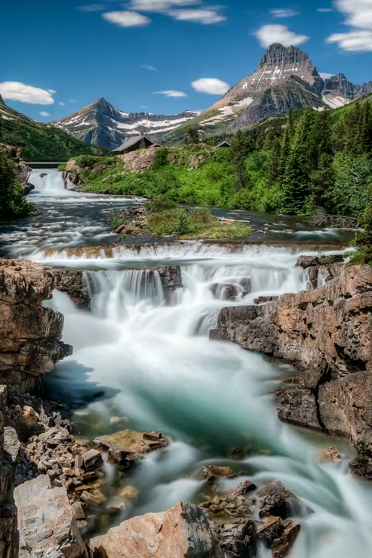 Workshop location; A long exposure creates silky, smooth water and streaking, puffy clouds surrounded by blue sky in this vertical composition of Swiftcurrent Falls in Glacier National Park, MT. The powerful river cascades through the scene initially surrounded by forest, then as it draws closer, warm hued rocks and cliffs bind the aqua blue water as it flows out the bottom right corner of the frame. Above, in the upper part of the image, two snow capped peaks stretch toward the sky.
