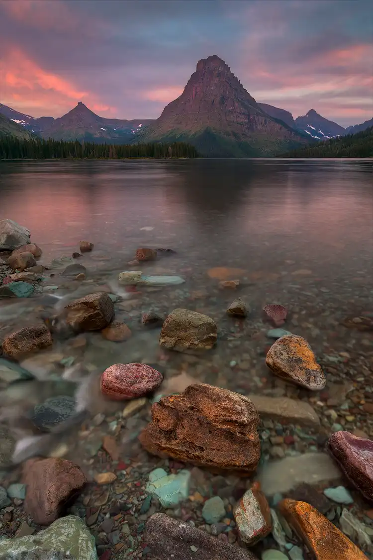 Photography location; The sky was a mix of clouds that had been bluish gray. Now, the first light of day catches the lowest hanging and they begin to glow a pinkish orange revealing their linear and curving geometry. Thrust into these clouds are three pyramid shaped peaks perched high above Two Medicine Lake in Glacier National Park. Scanning down this vertical image one sees the color of the clouds dancing in the rippled lake water and stretching toward the viewer and to the collection of partially submerged, colorful, red, green and orange rocks of the shoreline.