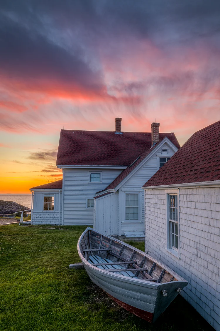 Workshop location; The sky is ablaze with clouds and sunset colors in the top third of this vertical image. The bottom portion of the image is filled with an old, lighthouse keepers house whose walls are a mix of ship lap and shake siding painted white and the roof is red shingles. The closest foreground element is an old row boat sitting along side the house.