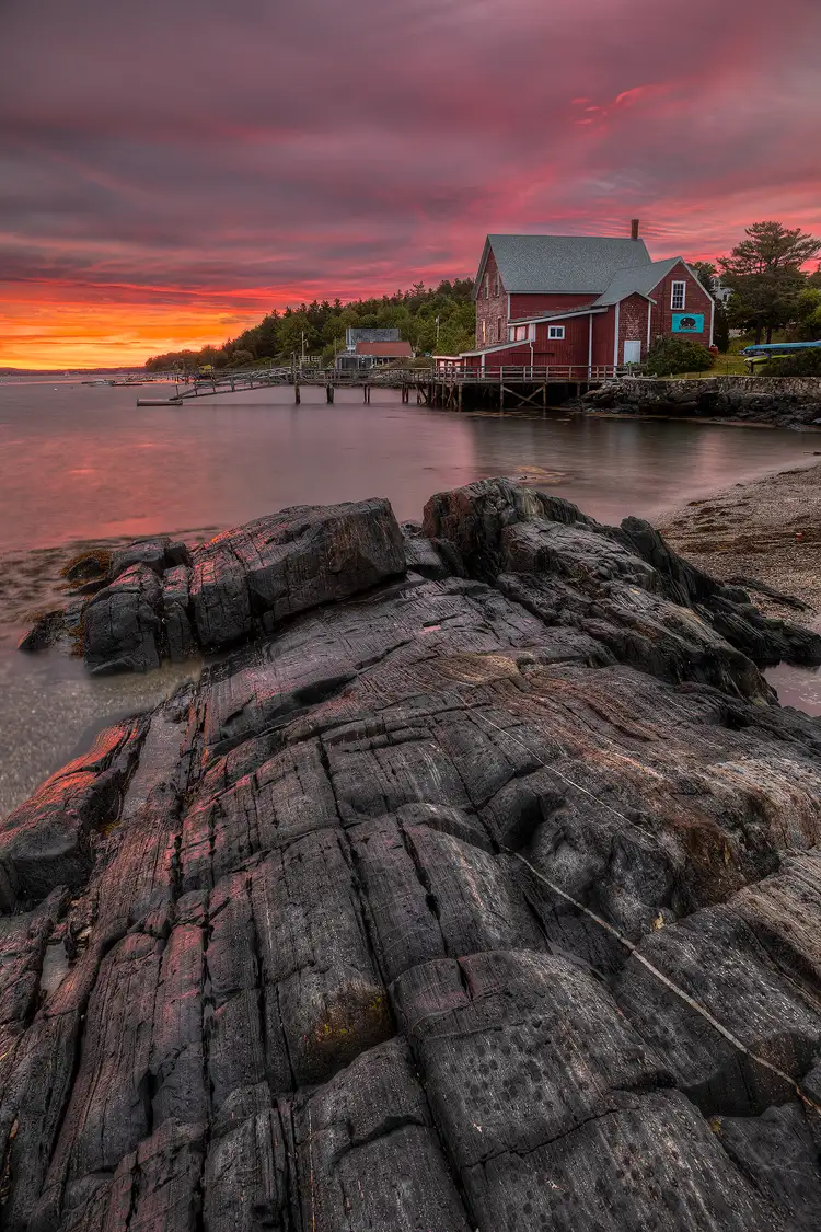 Workshop location; Vertical Maine coast image of striated black rock entering the bottom of the frame and leading up to the shoreline. A short distance away a red building is dockside and a fiery sunrise sky is above.