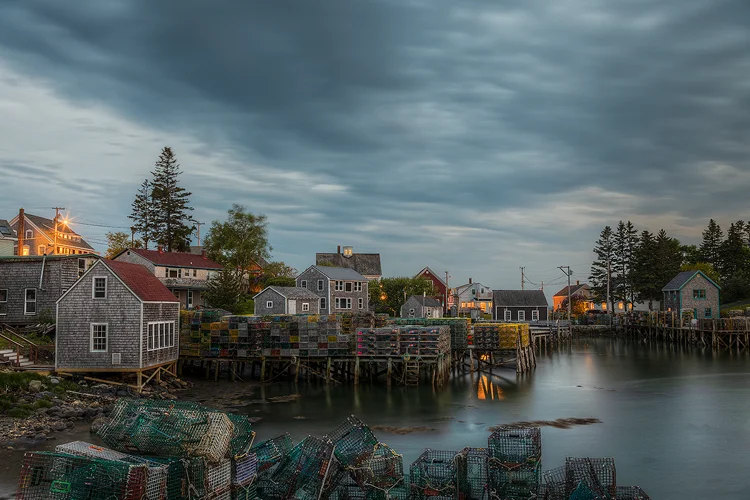 Workshop location; The sun has gone down and there is a slight, bluish feel to the image. The scene is a side view of the docks in Port Clyde, Maine, which are packed with lobster traps. Above the docks the small hamlet rises on a hill. Warm lights are beginning to glow in a few of the homes.