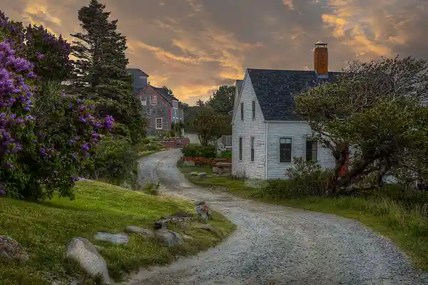 A narrow, dirt lane enters the bottom right of the frame and serpentines to the center of the image. This country lane is lined by green grass, blooming lilacs, the occasional tree and a few coastal style homes hugging close to the lane. The clouds above reflect various, sunset hues. These charming, quaint scenes offer a glimpse to another time and place and evoke emotions that compel many photographers to seek workshops on the Maine Coast.