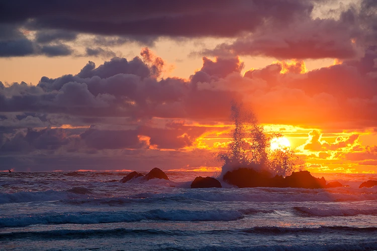 Photography location; Sunset photo of waves crashing on rocks at Cannon Beach on the coast of Oregon. The sun is visible behind splashes that are spiking into the air after a wave crashes on rocks. The sky is purple and orange and filled with clouds partially outlined by edge glow from back lighting.