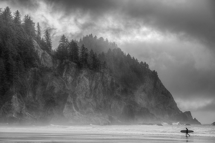 Workshop location; This is a long lens image which compresses a lone surfer exiting the water against a background of rugged, tree topped cliffs embroiled with stormy clouds.