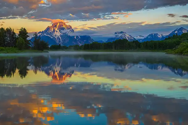 Warm, morning light illuminates clouds and the snow capped peak of Mount Moran. Far below, Mount Moran reflects in the calm, drifting water of Oxbow Bend in the Snake River. The sharply rising mountains of Grand Teton National Park create dramatic photographs that keep participants attending workshops here.