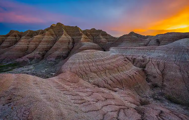 Eroded ridges leading toward layered buttes beneath a glowing sunset in Badlands National Park.