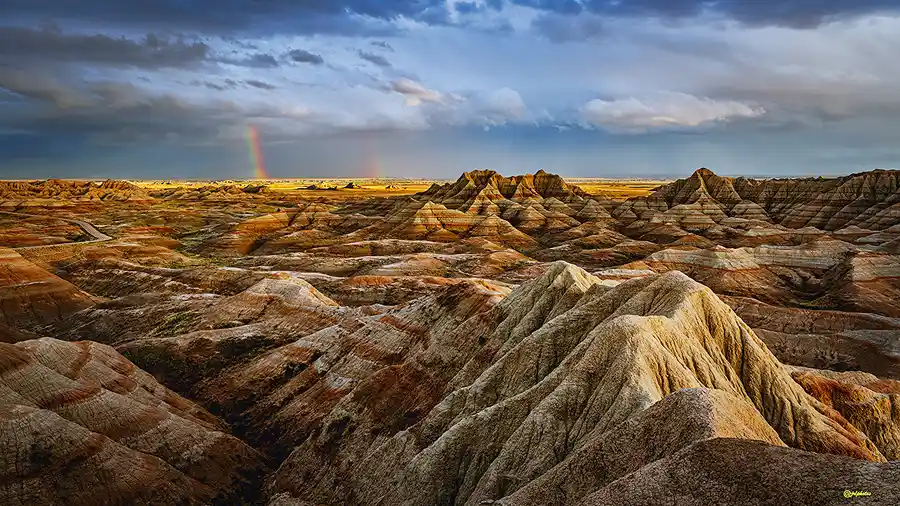 Evening light breaking beneath storm clouds to illuminate the layered badlands, with a faint rainbow on the horizon in Badlands National Park.