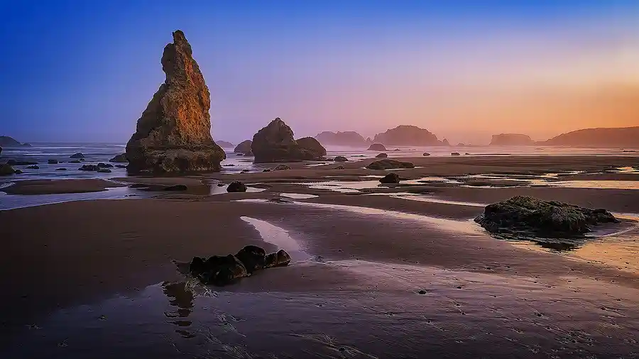 Panoramic sunset photo of Bandon Beach with glowing tide pools and sea stacks, including a prominent rock formation lit by warm evening light.