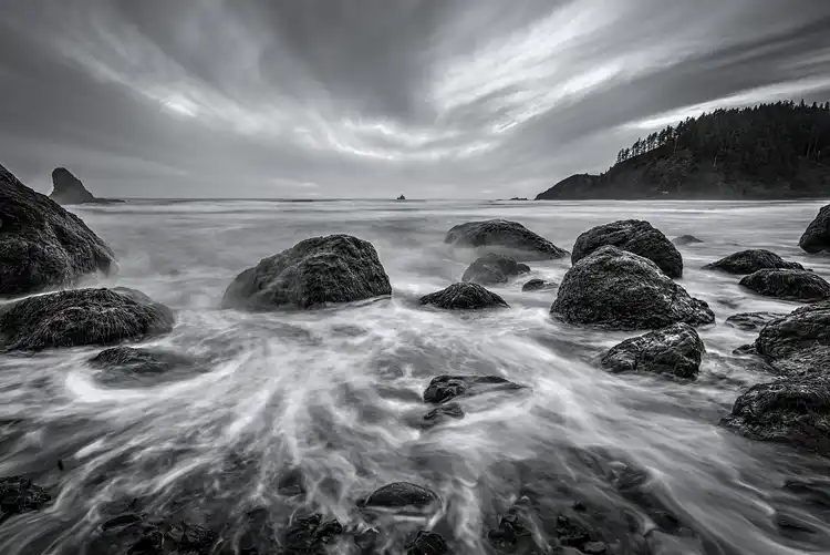 Long-exposure surf flowing around shoreline rocks beneath a dramatic sky on the Oregon coast.