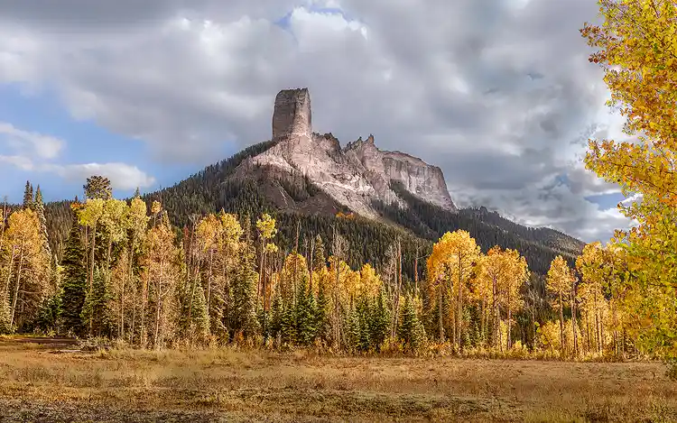 Chimney Rock rising above golden aspens beneath dramatic clouds in the San Juan Mountains of Colorado.