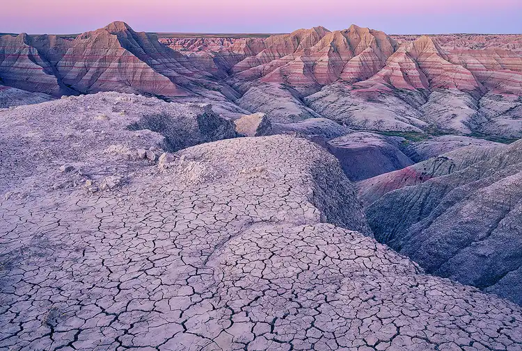 Pastel evening light lingering over the layered badlands beyond a cracked clay overlook in Badlands National Park.