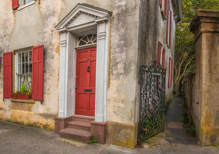 Weathered Charleston home with a red door beside a narrow alley disappearing between historic buildings.