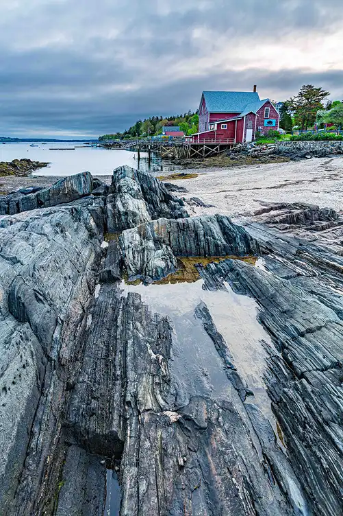 Grooved granite shoreline leading to a weathered red fishing building along the Maine coast.