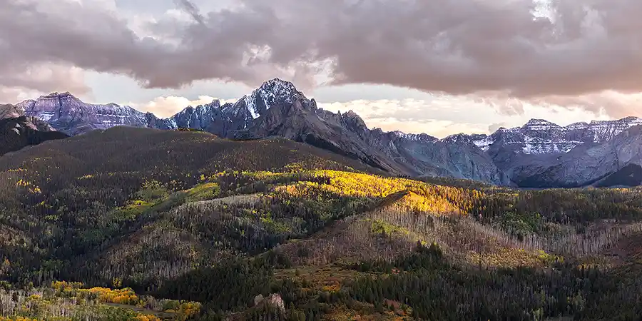 Evening light illuminating autumn hills beneath rugged peaks in the San Juan Mountains of Colorado.