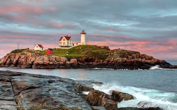 Waves crashing against the granite shoreline below Nubble Lighthouse beneath pink sunrise clouds on the Maine coast.