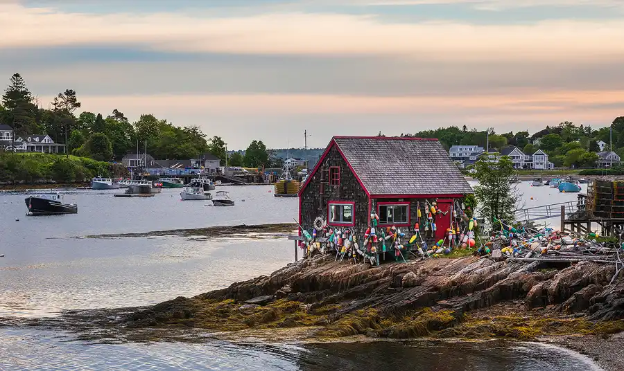 Colorful lobster buoys surround a weathered red trimmed fishing shack overlooking a quiet harbor on the Maine coast.