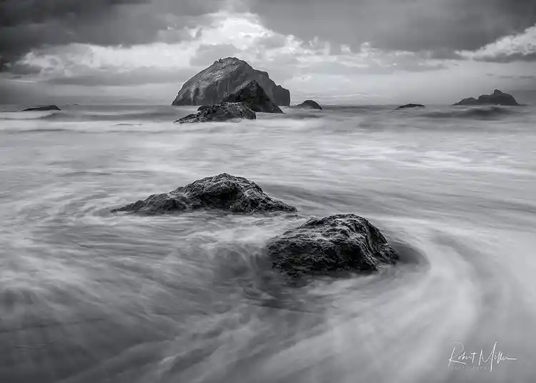 Long-exposure waves swirling around shoreline rocks with sea stacks beneath storm clouds on the Oregon coast.