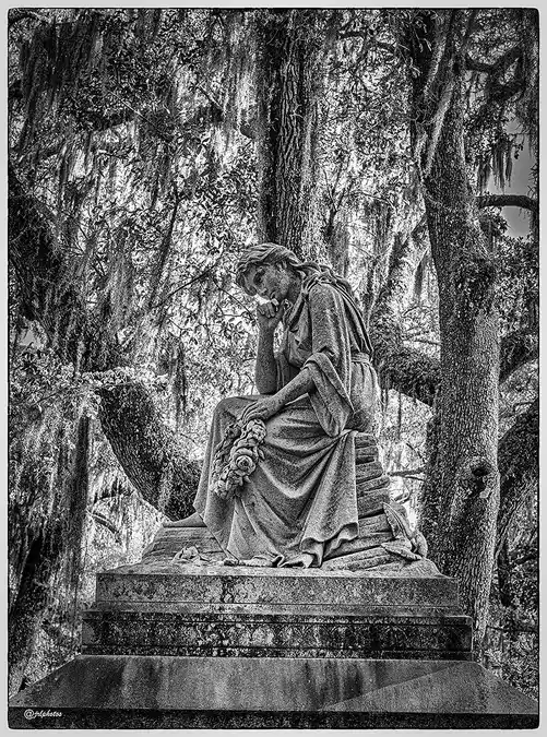 Seated stone memorial with bowed head beneath moss-draped live oaks in Bonaventure Cemetery.
