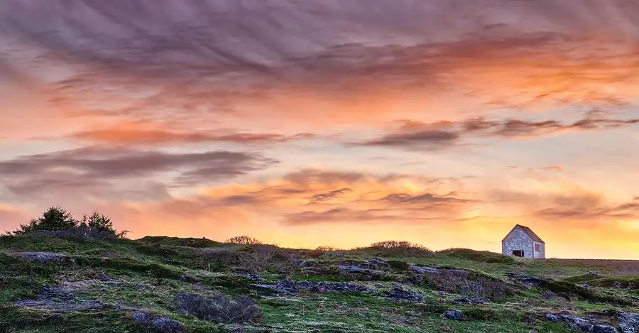 Historic barn standing alone beneath glowing sunset clouds on Manana Island off the Maine coast.