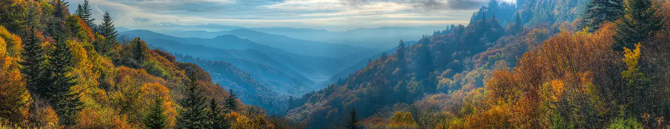 Workshop location; This high elevation, panoramic, image has back lit, autumn colors glowing on each side of the image forming a v shape that frames the view down a long twisting valley. The valley, in the middle of the frame, has alternating hazy, blue layered mountains dipping into it from each side repeating into the distance.
