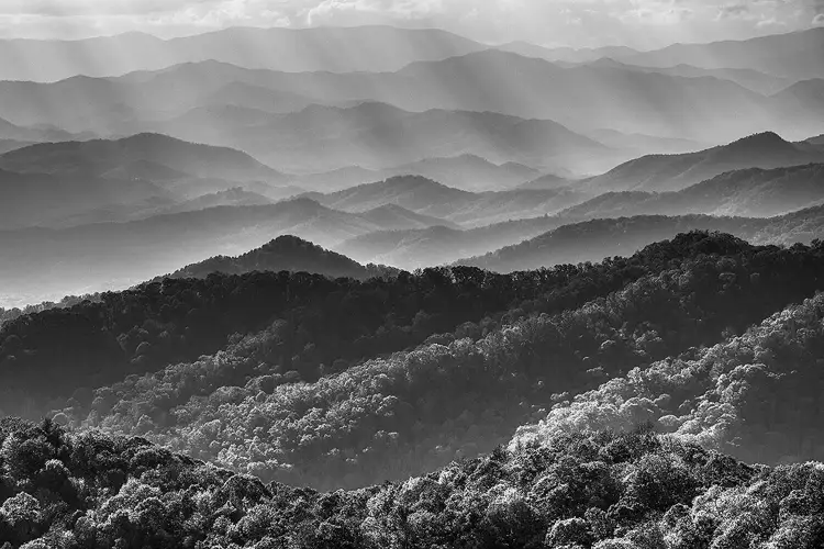 Workshop location; From a high view point, this is a long lens, black and white photo showing numerous ridge lines, layered mountains, through all of the image except for a small amount of hazy, partly cloudy sky at the top. It is hazy and cloudy enough to cause sun beams to project down into the top half of the image.