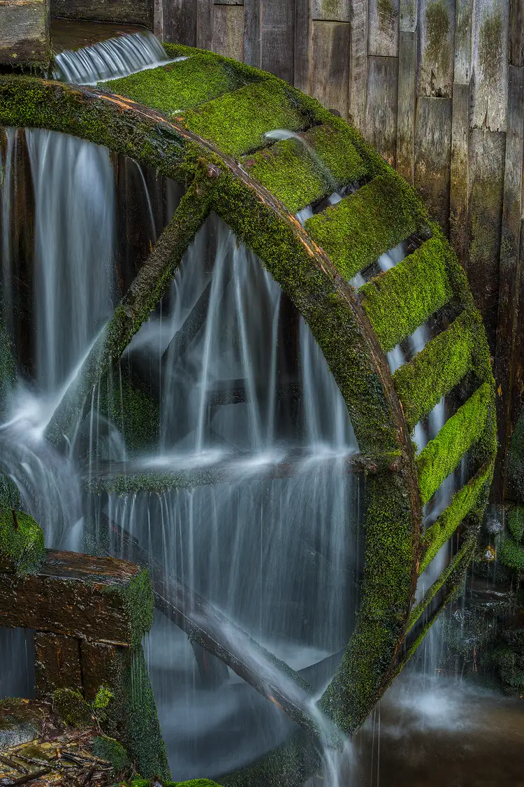 Photography location; Close-up, vertical image of the moss covered water wheel on Cable Mill in Cades Cove, Great Smoky Mountains National Park. The wheel is not spinning, but water spills through it and a long exposure makes the water record as silky strands.