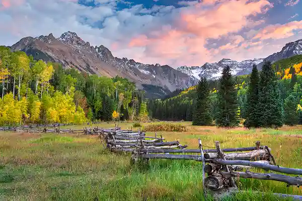 Photograph of an old, zig zag fence leading through an autumn field to a colorful stand of aspen trees with Mt. Sneffels catching sunset light towering above. Photographs like this make Colorado in the autumn on of the most sought after destinations for photography workshops.