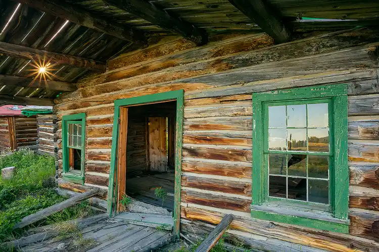 Workshop location; A dilapidated log cabin with a roof over hanging the front porch fills the frame, In the upper left corner the setting sun finds a small crack in the roof and a starburst records in the image.