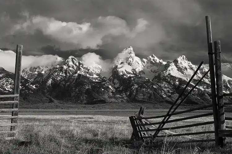 Photography location; Black and white photograph of a Mormon Row fence gate framing a stormy, cloudy sky swirling around mountains laden with fresh snow. Grand Teton National Park.