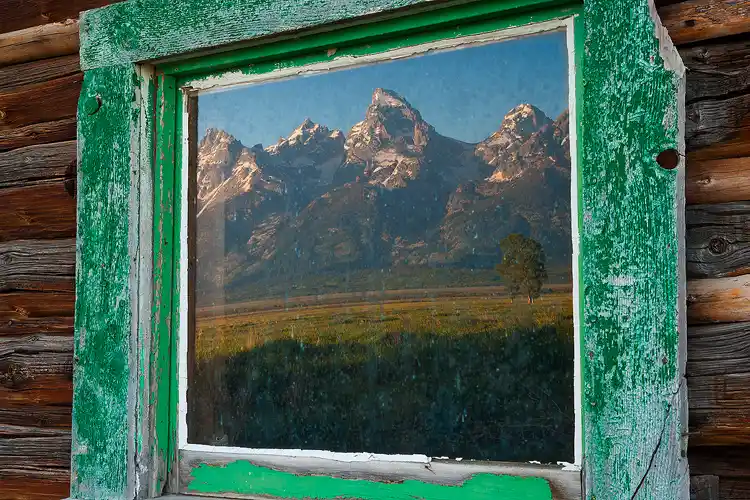 Photo of rugged mountains reflecting in the window of an old cabin in Grand Teton National Park, Wyoming.